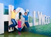 Three people—a man, a woman, and a young girl—jump in front of large, white "HOLLYWOOD" letters displayed against a blue and green backdrop.