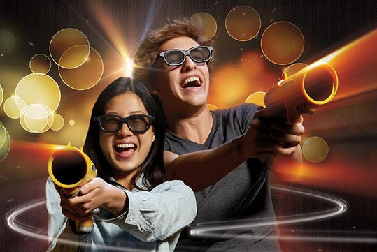Two people wearing 3D glasses smile and aim toy guns, with bright abstract lights and circles in the background.