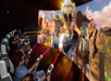 People seated in a theater use handheld controllers to interact with a large, curved screen displaying animated robots in a Wild West setting, with colored beams connecting them to the screen.