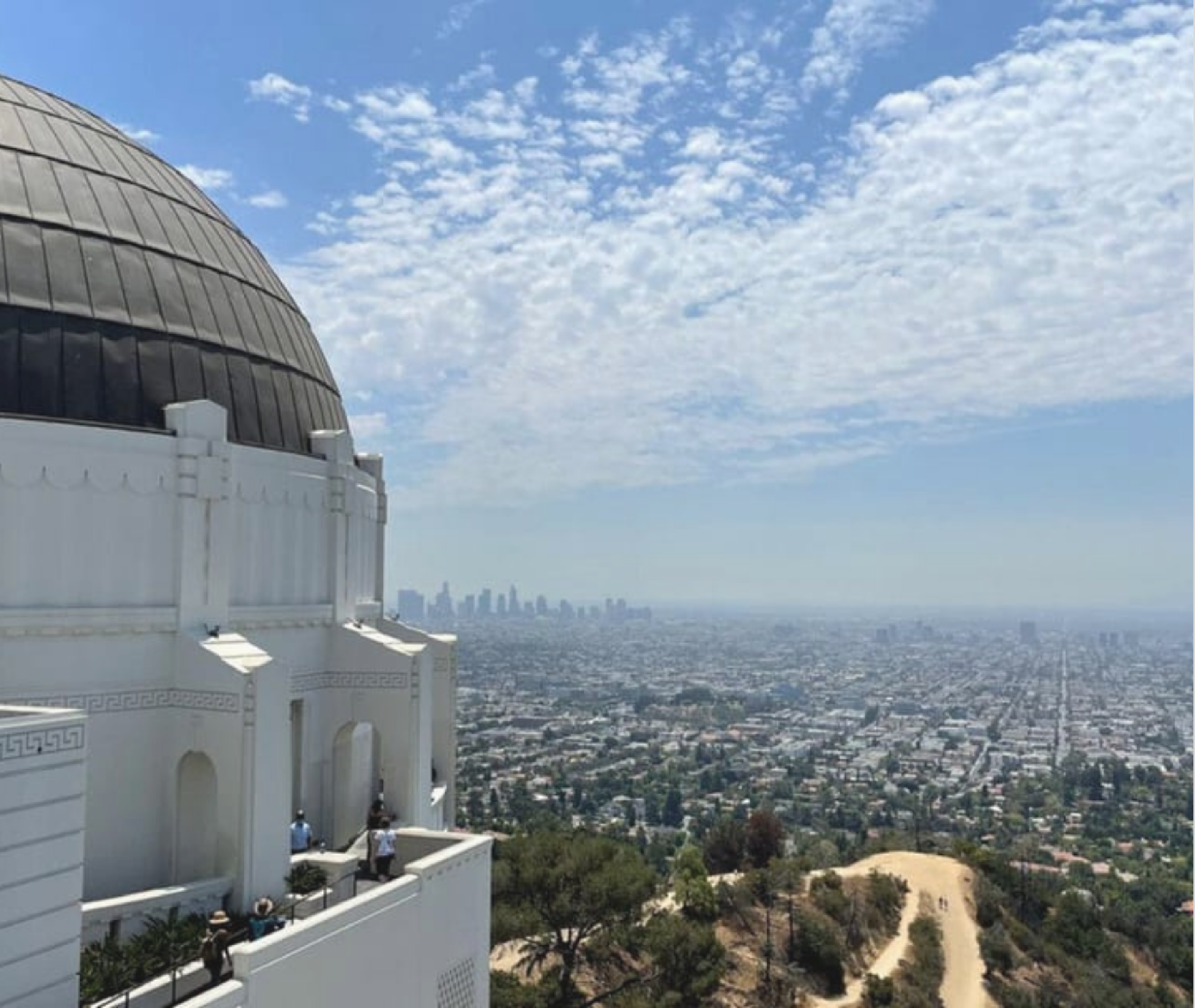 The Griffith Observatory is in the foreground on the left, overlooking a sprawling cityscape of Los Angeles under a partly cloudy sky.