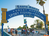 People walk under the iconic blue and yellow Santa Monica Pier entrance sign reading “Yacht Harbor, Sport Fishing, Boating, Cafes” on a sunny day with palm trees in the background.