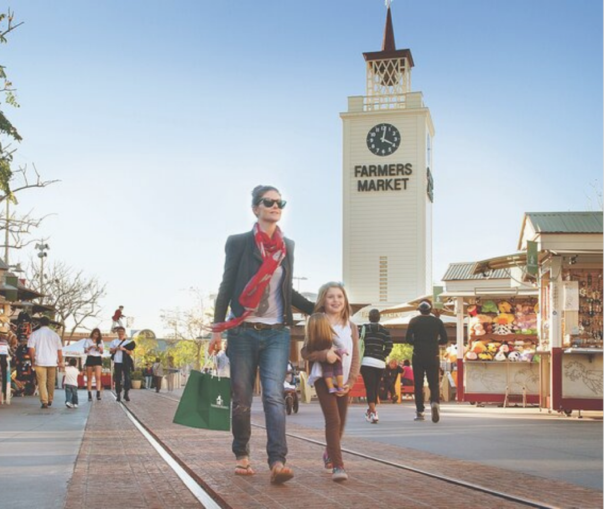 A woman and a young girl walk along train tracks near outdoor stalls, with a tall Farmers Market clock tower in the background.