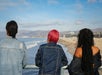 Three people with different hairstyles stand by a railing, looking out at the ocean and sandy beach with mountains in the distance under a partly cloudy sky.