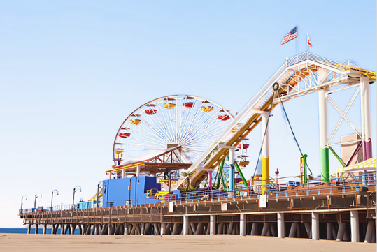 A large ferris wheel and roller coaster are on a pier with flags on top, set against a clear blue sky and sandy beach.