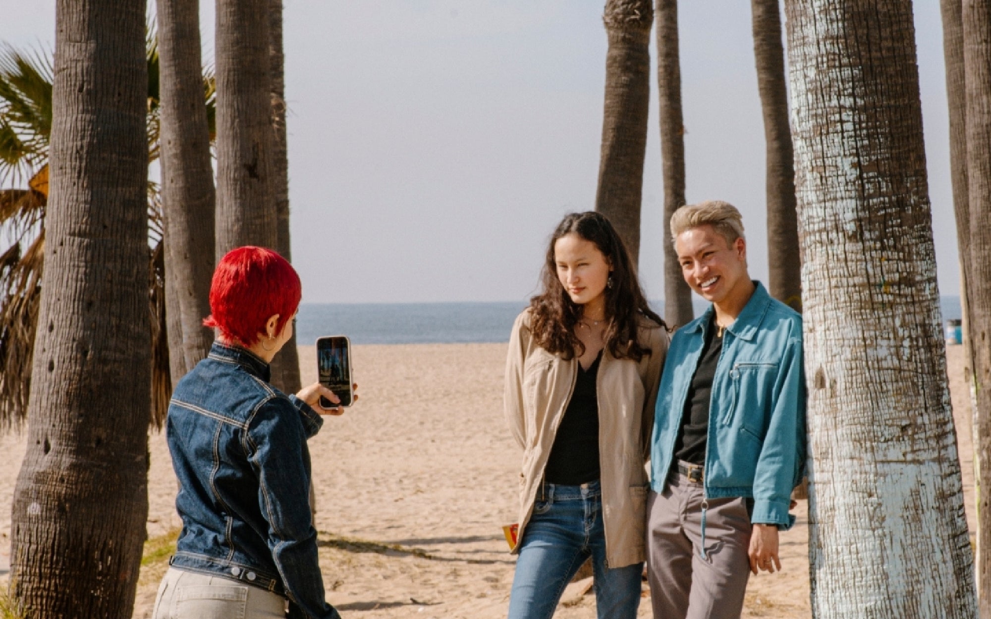 A person with red hair takes a photo of two people posing and smiling on a sandy beach with palm trees and the ocean in the background.