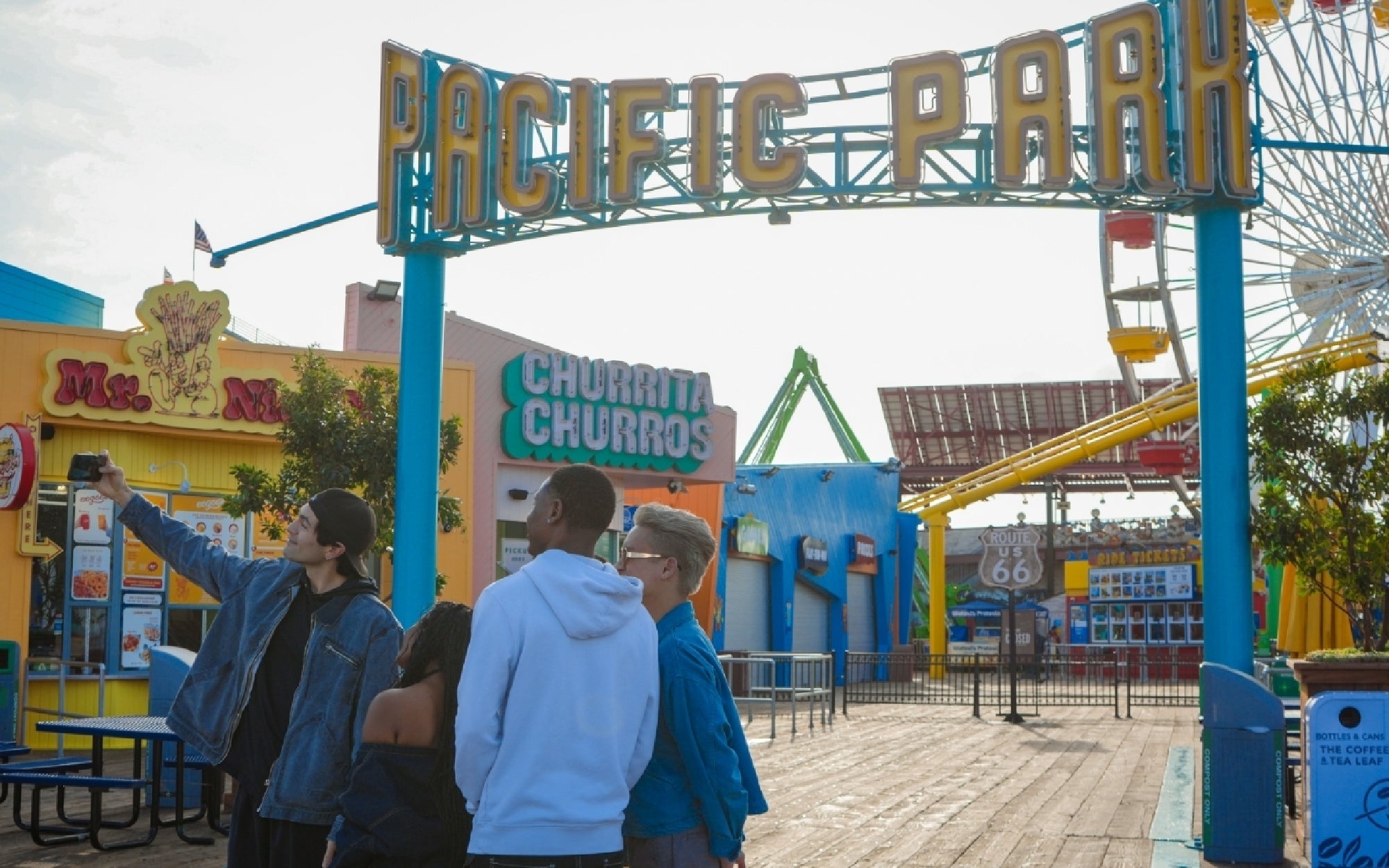 Four people take a selfie under the Pacific Park entrance sign on a wooden boardwalk, with food stands and amusement park rides visible in the background.