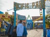Four people take a selfie under the Pacific Park entrance sign on a wooden boardwalk, with food stands and amusement park rides visible in the background.