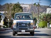 A tour van with several passengers drives on a street in Los Angeles, with the Hollywood sign visible on the hillside in the background.