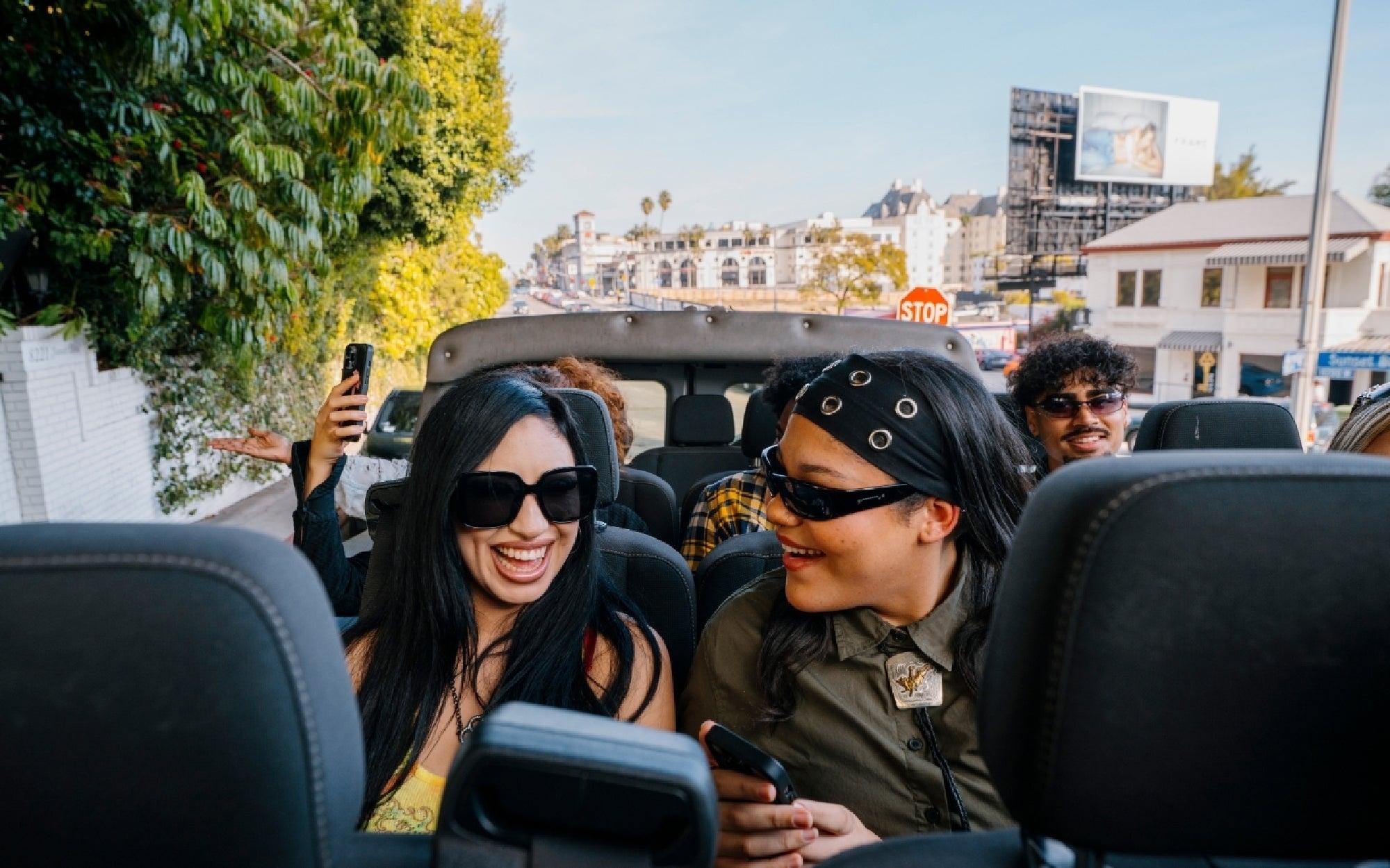 A group of people ride in a convertible car with the top down, smiling and taking photos on a sunny day in the city.