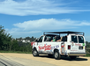 An open-air Starline Tours van with passengers is parked on a roadside, overlooking a scenic area with trees and distant houses under a blue sky.