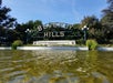 A low-angle view of the Beverly Hills sign above a shallow pool of water, with greenery and trees in the background under a clear blue sky.