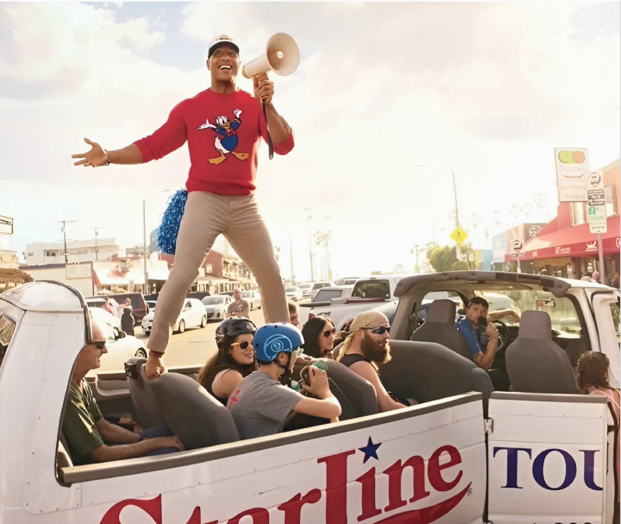 A person in a red shirt stands on the seat of a Starline tour bus holding a megaphone, while several passengers sit and look on in an outdoor urban setting.