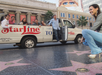 A group of people pose by a Starline Tours van while a woman crouches near Hollywood Walk of Fame stars on a sunny day.