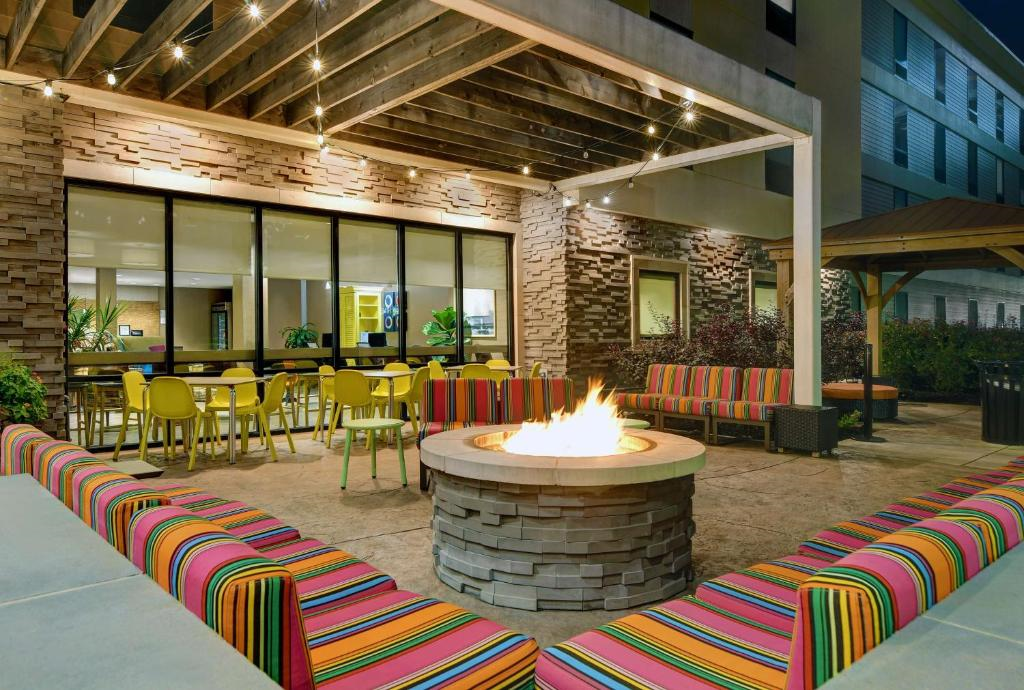 Outdoor patio with colorful striped seating around a circular fire pit. Tables and chairs are arranged near a building with large windows. String lights hang from a wooden pergola.