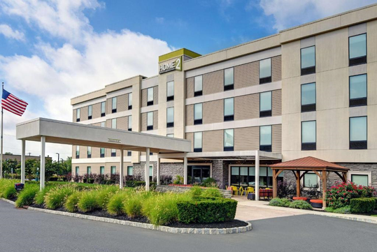Four-story hotel building with an American flag outside, featuring a covered entryway and landscaping with shrubs.
