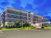 Four-story hotel building with a modern design, featuring an entrance canopy. An American flag is visible, and parking spaces are in front. The sky is a vibrant blue.