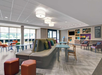 Modern hotel lobby with colorful seating, tables, and a snack area. Large windows provide natural light.