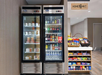 Two beverage refrigerators filled with drinks and snacks next to a shelf of candy and chips in a market corner under a "Home2 MKT" sign.
