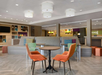 A modern hotel lobby with a seating area featuring orange chairs, a round table, and a large gray bench. The space includes ambient lighting and shelves with decorative items.