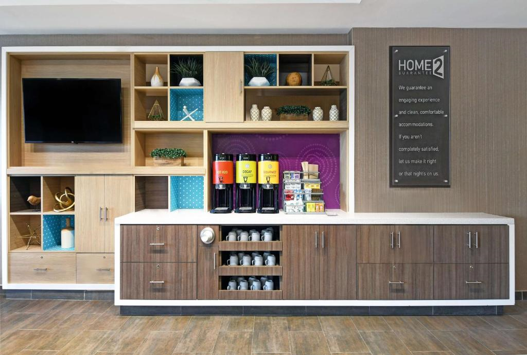 A modern beverage station with drink dispensers, cups, and a variety of decor items on wooden shelves. A large TV is mounted on the left.