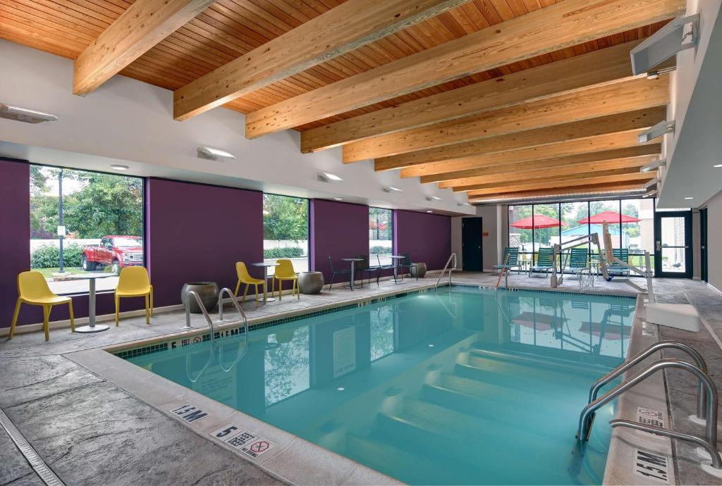 Indoor pool with wooden beams, surrounded by purple walls, yellow chairs, and small tables. Large windows provide natural light, with a view of outdoor umbrellas and trees.