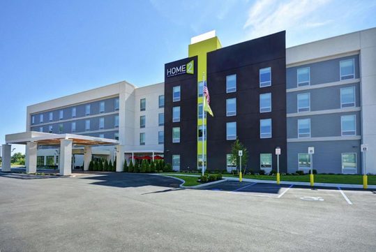 Exterior view of a modern, multi-story hotel with a large sign reading "Home2" on the facade, featuring a canopy entrance and surrounding parking lot.