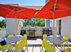 Outdoor patio with red umbrellas, yellow chairs, and tables. Two grills are set up in the background against a wall.
