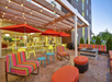 Outdoor patio with colorful furniture, string lights, and red umbrellas. Large windows behind reveal an indoor seating area with vibrant chairs.