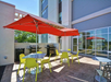 Outdoor patio with red umbrellas, yellow chairs, and tables. Two grills are positioned near the seating area, against a modern building backdrop.