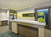 Hotel reception area with a modern design, featuring a front desk, shelves with snacks and drinks, a bowl of green apples, and a sign that reads "Home2.