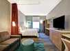 A modern hotel room with a bed, sofa, TV, desk, and a small round ottoman. The walls are decorated with patterned wallpaper and a framed picture. Natural light comes through a window.