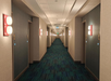A long hotel hallway with closed doors on both sides, featuring wall-mounted lights and a patterned carpet floor.