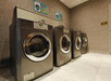 Four modern washing machines are lined up against a textured wall in a laundry room. Two signs are visible above the machines.