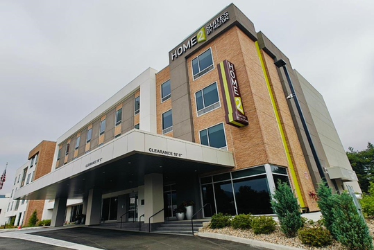 Four-story hotel with brick and beige exterior, labeled "Home2 Suites." Front entrance has a canopy, and a "clearance 11' 6''" sign. Small landscaped area with bushes in front.