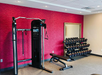 Gym with a red accent wall featuring a multi-gym machine, a set of dumbbells on a rack, and an adjustable bench on a carpeted floor.