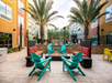 Outdoor patio with turquoise chairs around a central fire pit, surrounded by palm trees and colorful building facades.