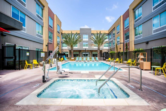 Outdoor pool and hot tub area at a modern hotel, surrounded by yellow chairs, with palm trees and building facade in the background under a clear sky.