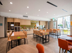 A spacious, modern breakfast area with wooden tables and chairs, a buffet counter labeled "inspired table," and natural light streaming through large windows.