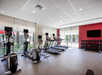 A gym with treadmills, exercise bikes, wall-mounted TVs, and a rack of dumbbells against a red wall. Large windows offer natural light.
