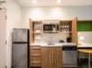 A compact kitchen with a fridge, microwave, sink, dishwasher, and shelves holding dishes and mugs. A small countertop workspace and a desk area with a lamp are adjacent.