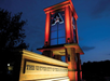 Tower with university signage and lighting, illuminated in red and blue at dusk. Forested area nearby.
