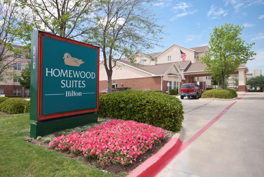 Entrance of Homewood Suites by Hilton with a sign, landscaped bushes, and pink flowers, along a driveway leading to the hotel's main building.