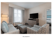 A modern hotel living room with a couch, chair, ottomans, TV on a stand, and a small window on the far left wall.