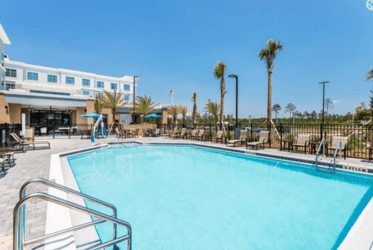 A bright blue outdoor swimming pool with lounge chairs lining the sides and palm trees in the landscaping on a sunny day.
