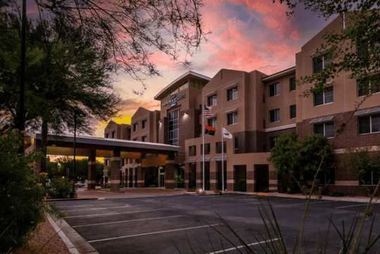 Modern hotel exterior with multiple stories and a covered entrance, surrounded by trees. Flags are flying near the entrance. The sky has a warm, colorful sunset.