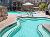 Outdoor pool area with a hot tub in the foreground, surrounded by patio chairs and tables. A larger swimming pool is visible in the background, bordered by a fenced area and building.