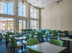 A modern lounge area with green and patterned chairs, tables, large windows, and ceiling fans. Natural light fills the space.