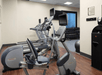 A gym room with elliptical machines, a stationary bike, and treadmills. Dumbbells are on racks in the background. The walls are beige, and the floor is wooden.