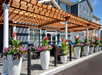 Outdoor seating area with a wooden pergola and large white planters filled with colorful flowers in front of a gray building.