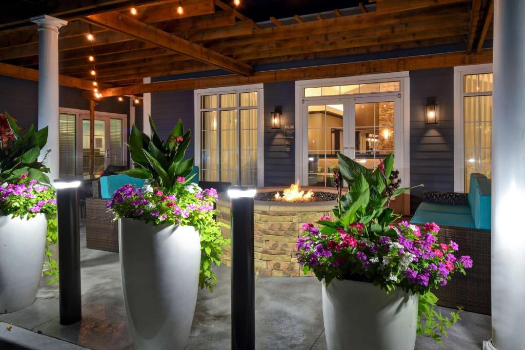 Outdoor patio with a fire pit, surrounded by wicker furniture and large planters with colorful flowers, under wooden pergola with string lights.